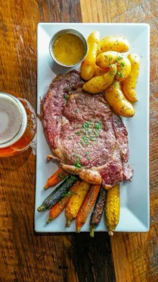 Overhead view: Steak, roasted potatoes, carrots, and beer on rustic wood.