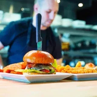 Gourmet burger held by a knife, with fries and blurred chef.