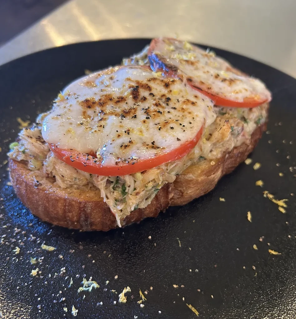 Toasted bread with seafood salad, melted cheese over tomato, on dark plate.