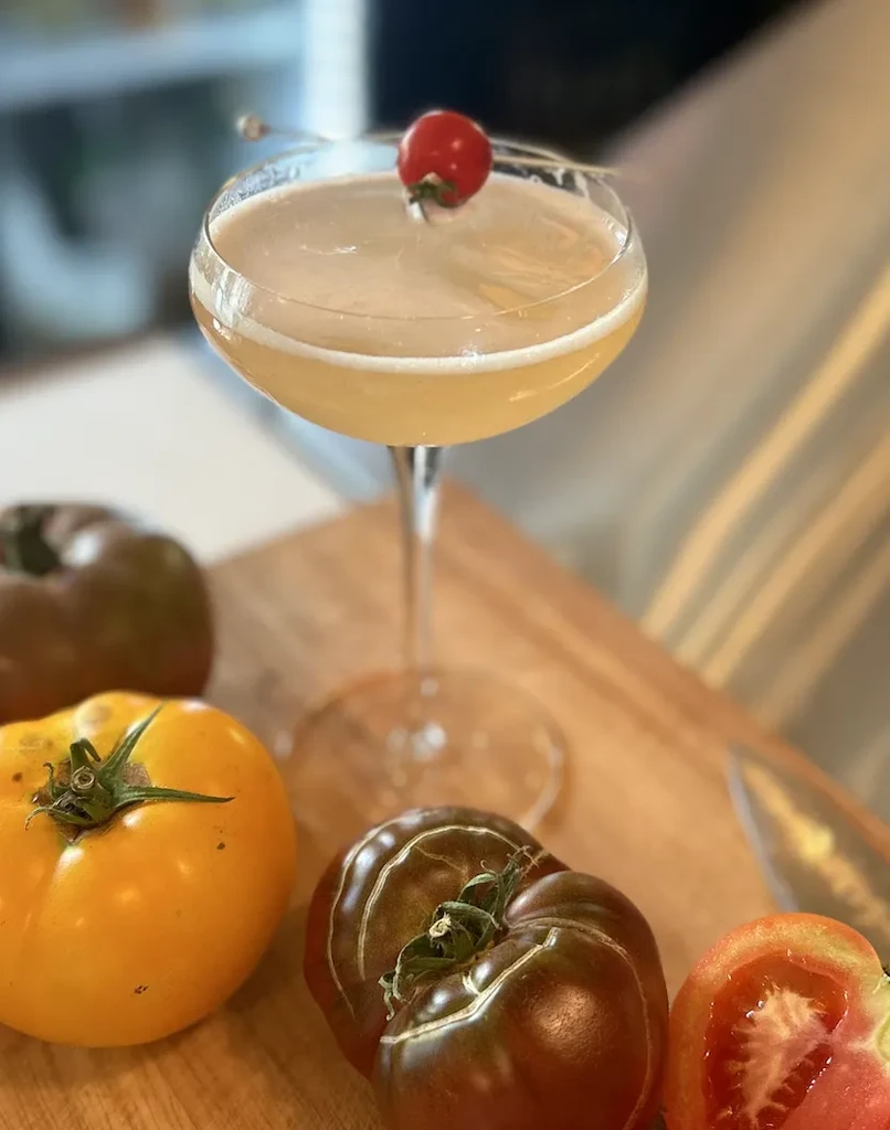 Cocktail in coupe glass with cherry, surrounded by colorful heirloom tomatoes.
