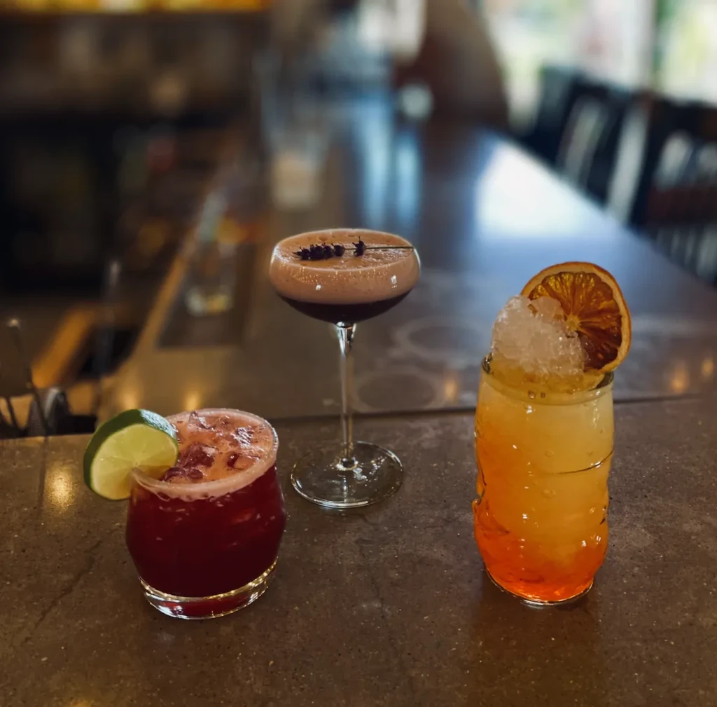 Three colorful cocktails with unique garnishes on a dim bar.