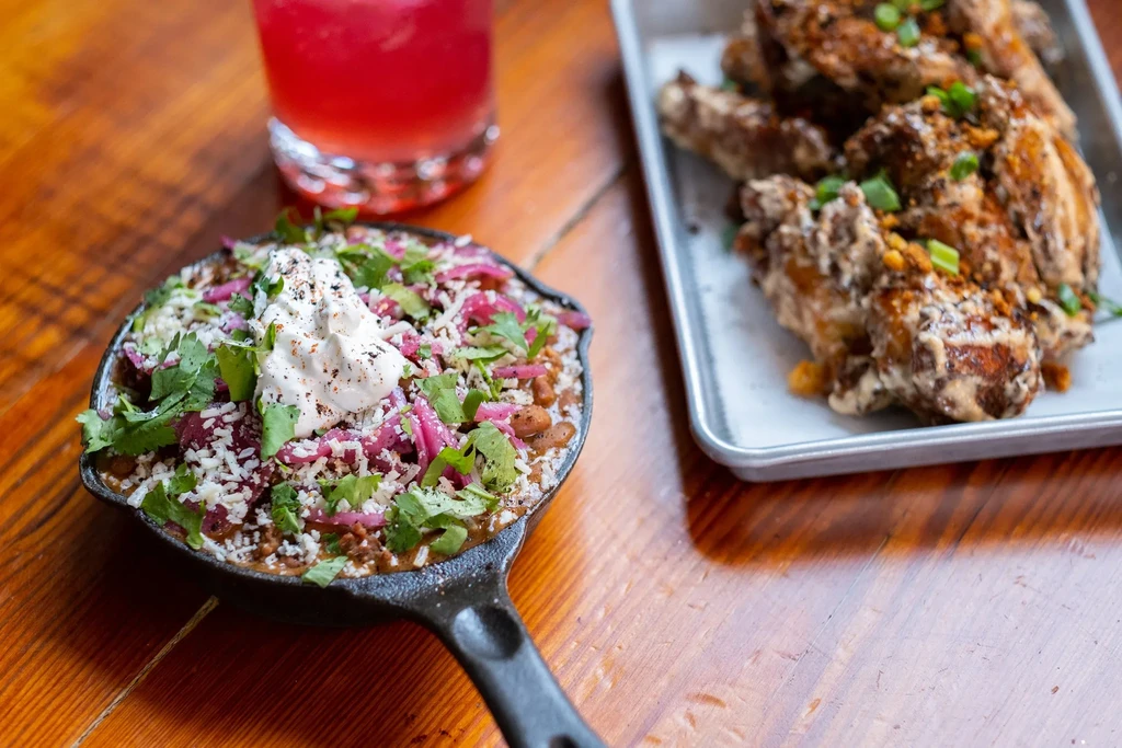 Close-up of chili skillet with toppings, blurred wings, and a red drink.