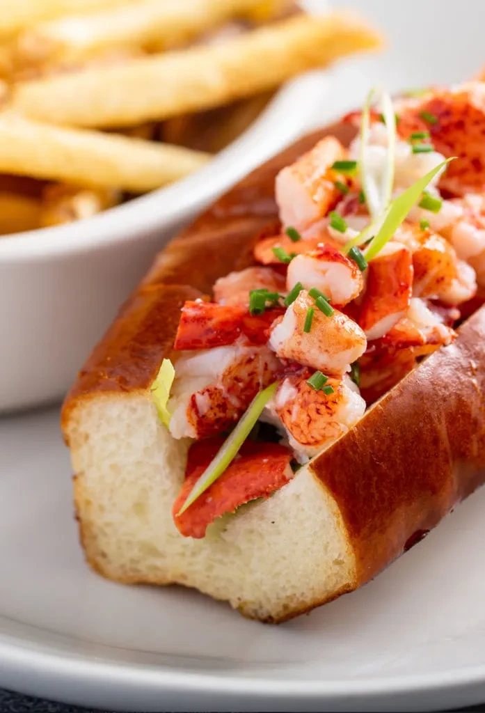 Toasted lobster roll filled with meat, garnished with chives and fries.