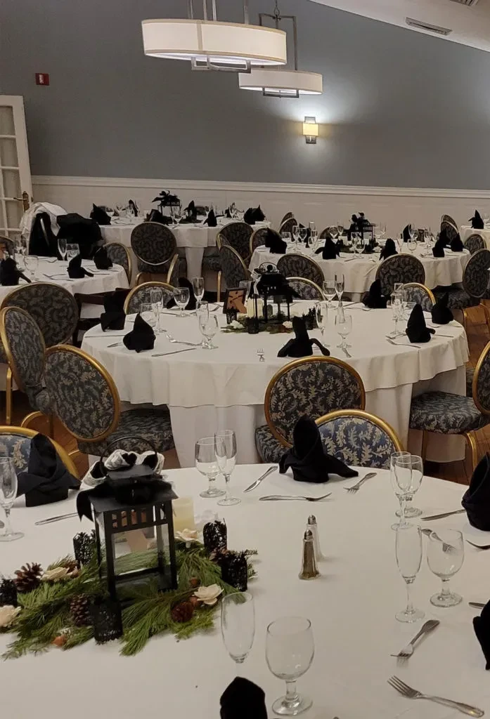 Formal event hall with round tables, lanterns, evergreen, and ornate chairs.
