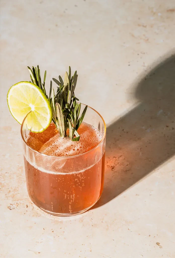 Bubbly amber drink in glass with lime and rosemary on stone.