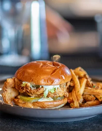 Close-up of fried chicken sandwich, lettuce, pickles, fries on dark plate.