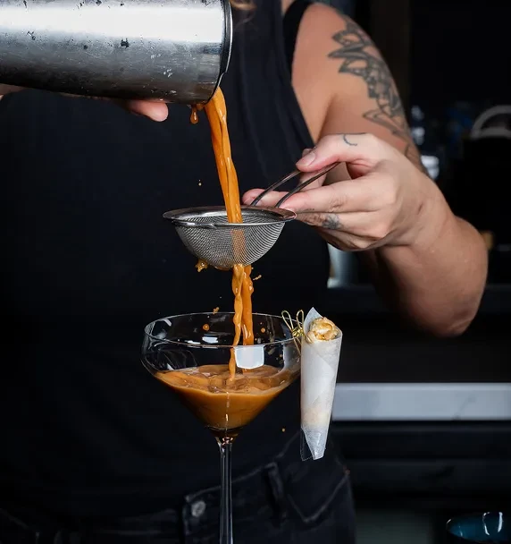 Tattooed person pours caramel cocktail through strainer into martini glass.