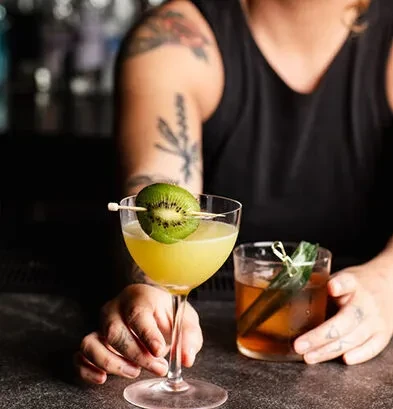 Yellow-green cocktail with kiwi, plus another, held by tattooed hands.