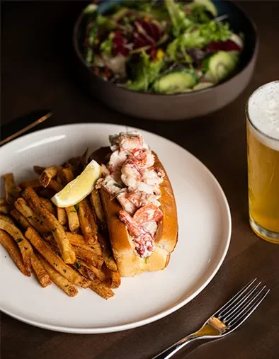 Lobster roll, french fries, beer, and salad served at a restaurant.
