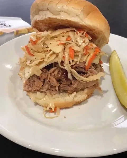 Pulled pork sandwich with coleslaw and a dill pickle on a white plate.