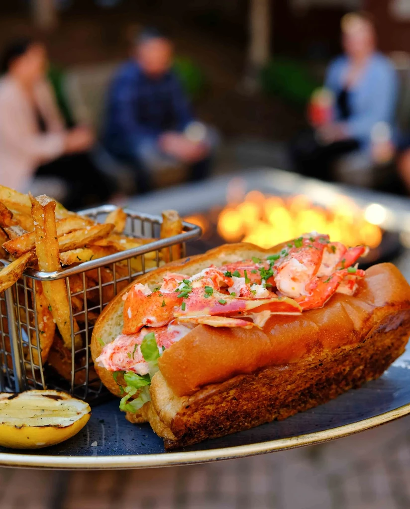 Close-up: Lobster roll, french fries, grilled lemon in outdoor setting.