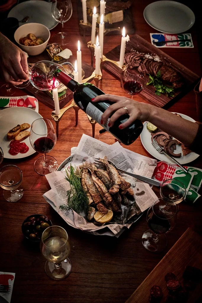 Candlelit rustic table with fried fish, meats, bread, wine, hands pouring.