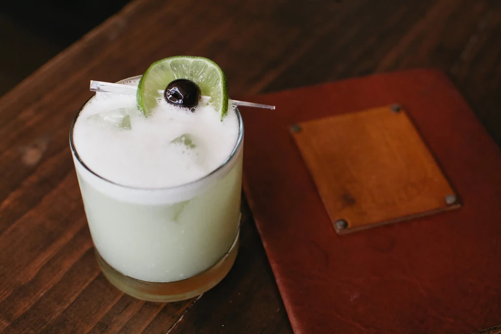 Pale green cocktail with white foam, lime, and cherry garnish.