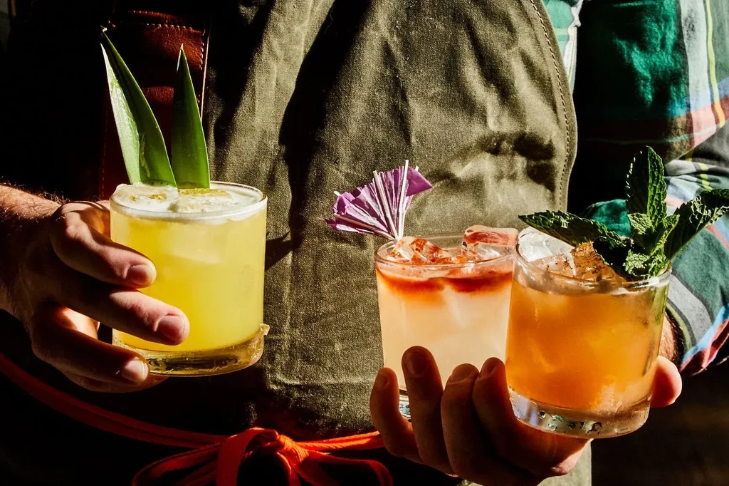 Three vibrant, garnished cocktails held by a person wearing an apron.