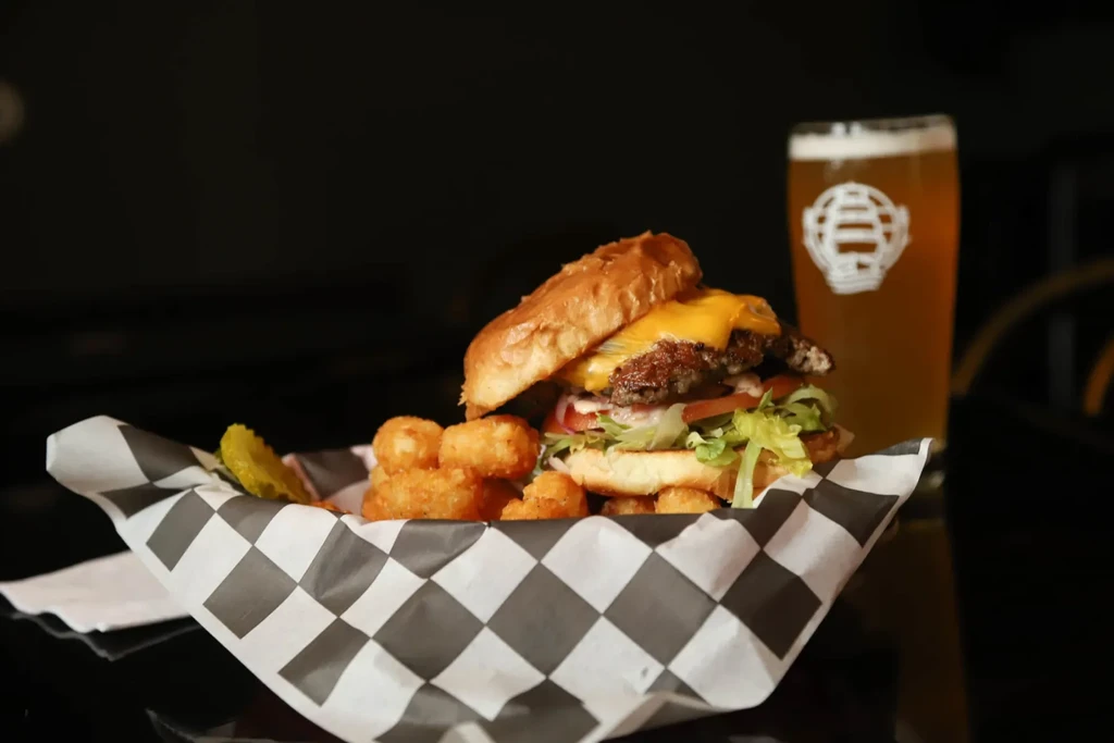 Cheeseburger, tater tots, pickle, and beer in a checkered basket.