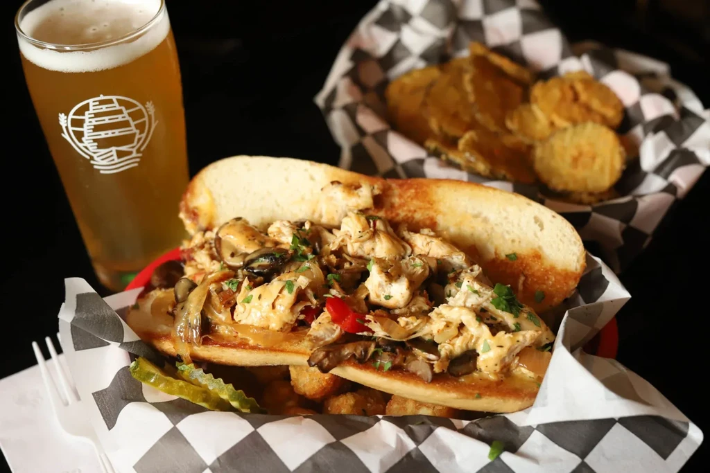 Large chicken and mussel sandwich, tots, and beer on a red tray.