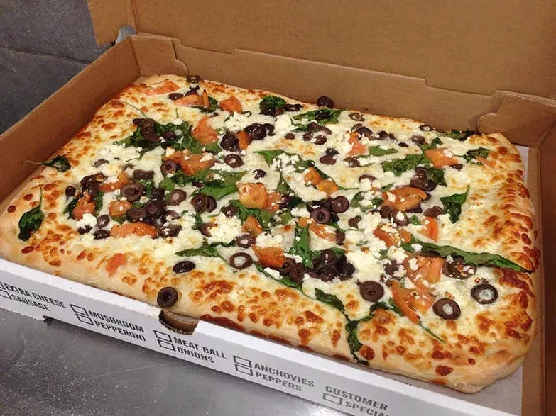 Rectangular pizza in a box, topped with cheese, olives, tomato, spinach.