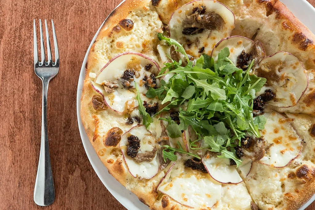 Freshly baked pizza with cheese, sliced fruit, dark figs, and arugula.