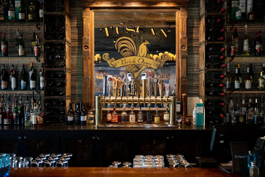 Brass beer taps, liquor bottles, and mirrored 'TUSCAN BRICK OVEN' sign.