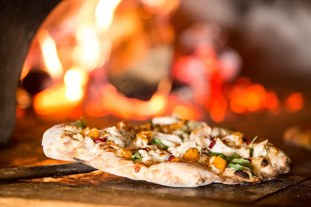 Thick-crust pizza with squash, chicken, and herbs near a wood-fired oven.