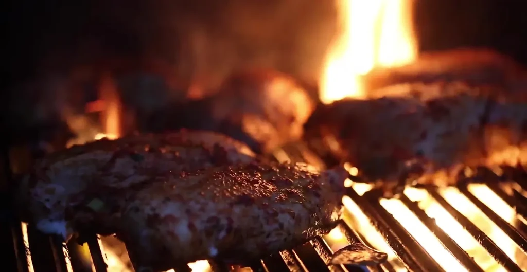 Close-up of seasoned meat grilling over bright orange flames.