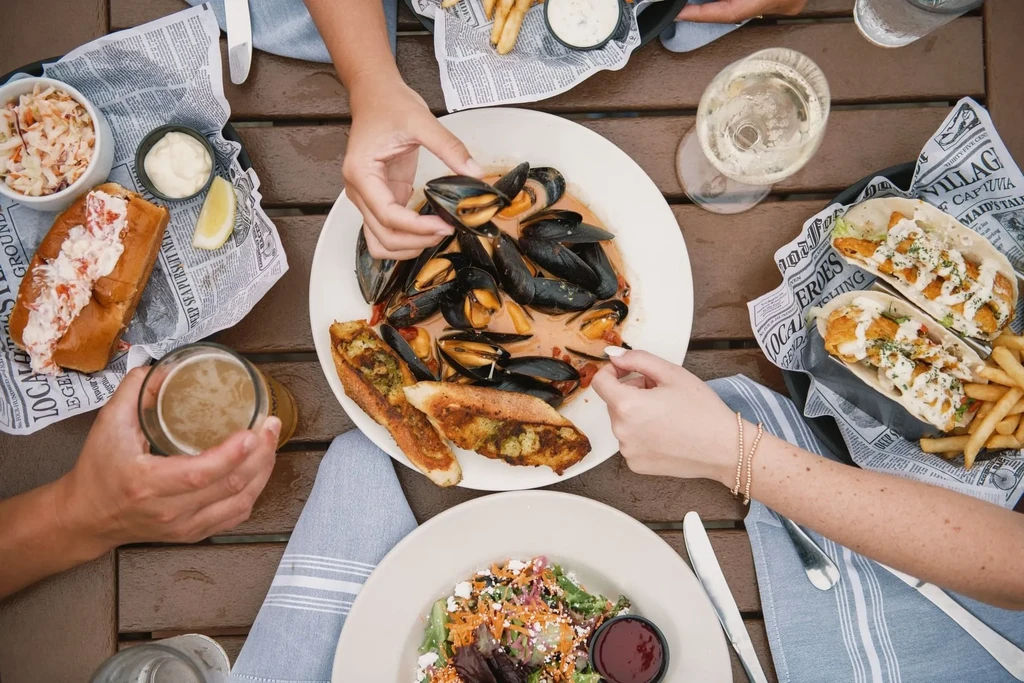 Overhead view: Multiple hands sharing seafood, fries, and drinks outdoors.