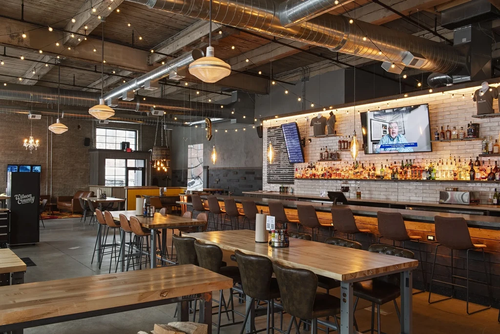 Empty modern industrial-rustic restaurant with bar, wood tables, and string lights.
