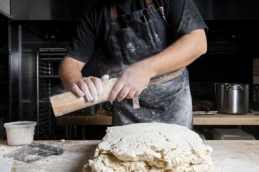 Baker's hands tap dough with a wooden block, flour in air.