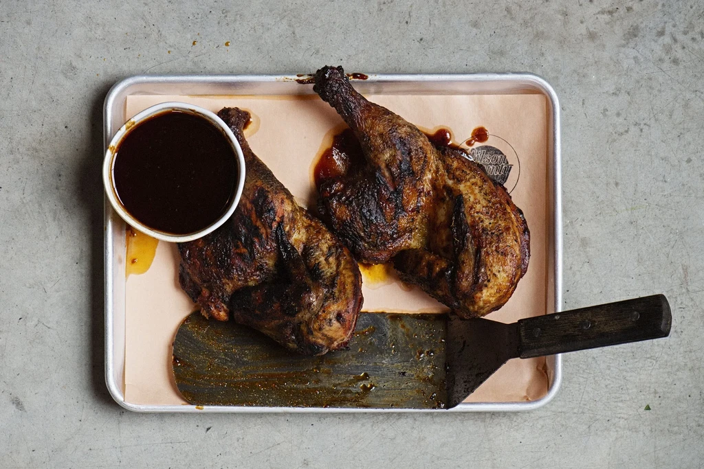 Grilled chicken halves on parchment-lined tray with sauce and spatula.