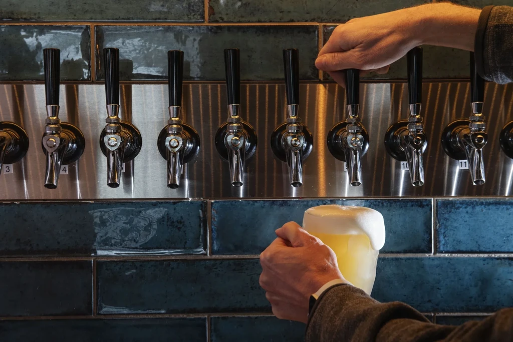 Hands pulling a chrome beer tap, holding an overflowing foamy mug.