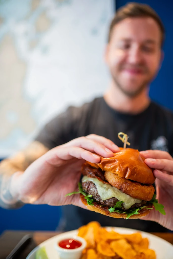Hands hold gourmet burger with onion rings, cheese, fries, and ketchup.