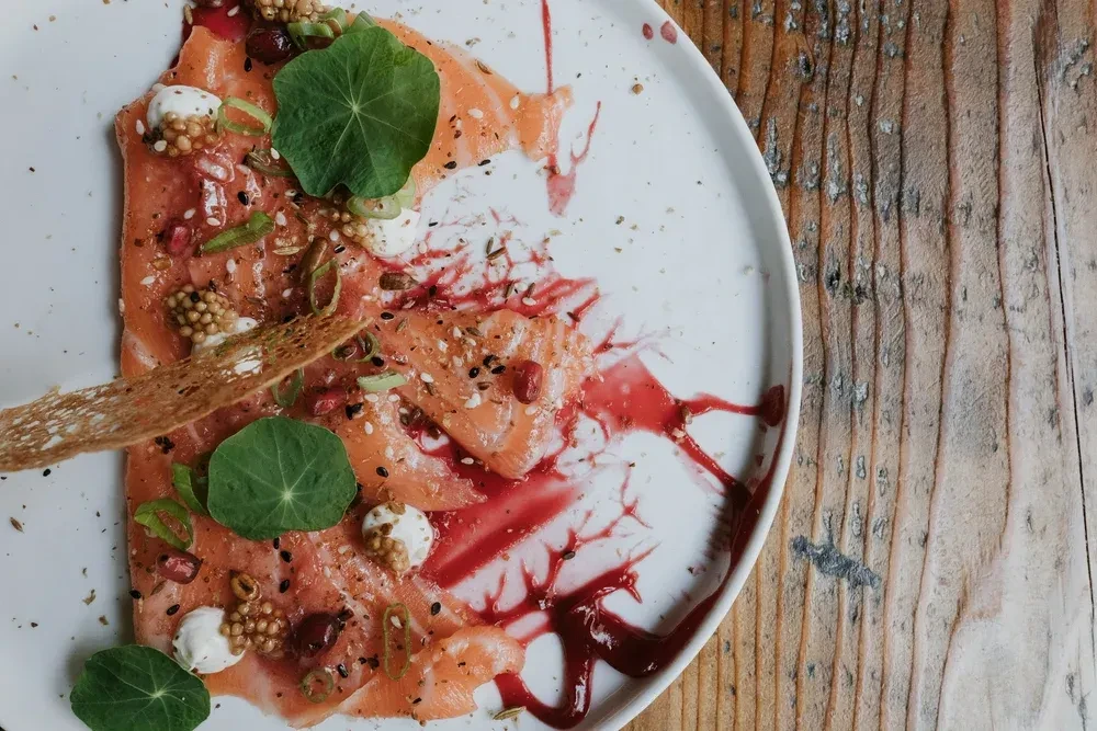Thinly sliced salmon carpaccio garnished with seeds, leaves, sauces, and cracker.