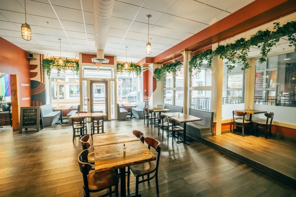 Empty modern restaurant interior with tables, booths, plants, and a rainbow flag.