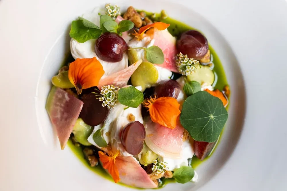 Plated dish: creamy cheese, grapes, radish, greens, flowers, green sauce.