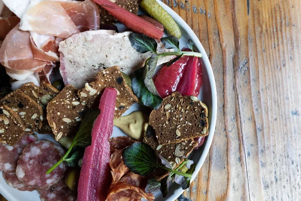 Overhead charcuterie board with cured meats, crackers, pickled vegetables, herbs, and mustard.