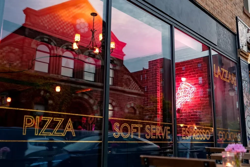 Lazzari restaurant facade with neon signs, offerings, and city reflections.
