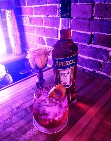 Cocktail, Aperol, and rose on bar with dramatic purple light.