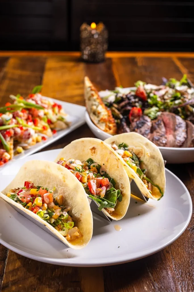 Three soft-shell tacos, steak salad, and appetizer on a wooden table.