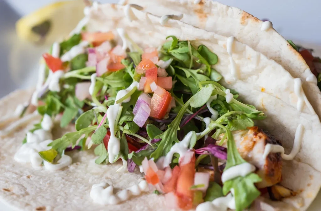 Close-up taco with greens, red tomatoes, purple onions, creamy sauce, protein.