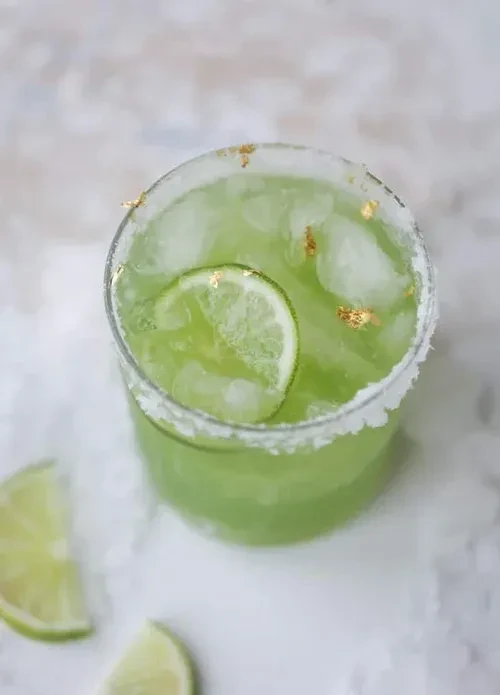 Green drink with ice, lime slice, sugar/salt rim, and gold flakes.