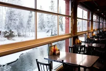 Empty restaurant tables overlook a snowy winter landscape through large windows.