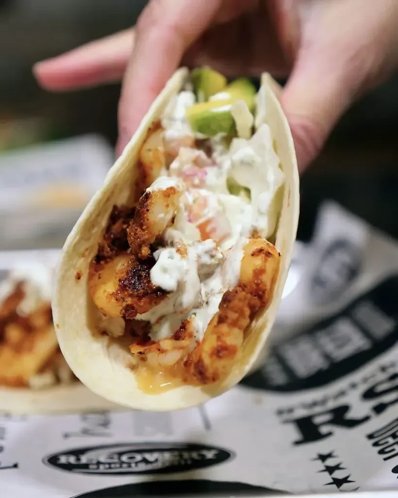 Hand holding a crispy shrimp taco with avocado, red salsa, and white sauce.