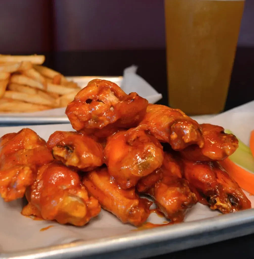 Plate of saucy buffalo chicken wings, celery, carrots, and blurred fries.