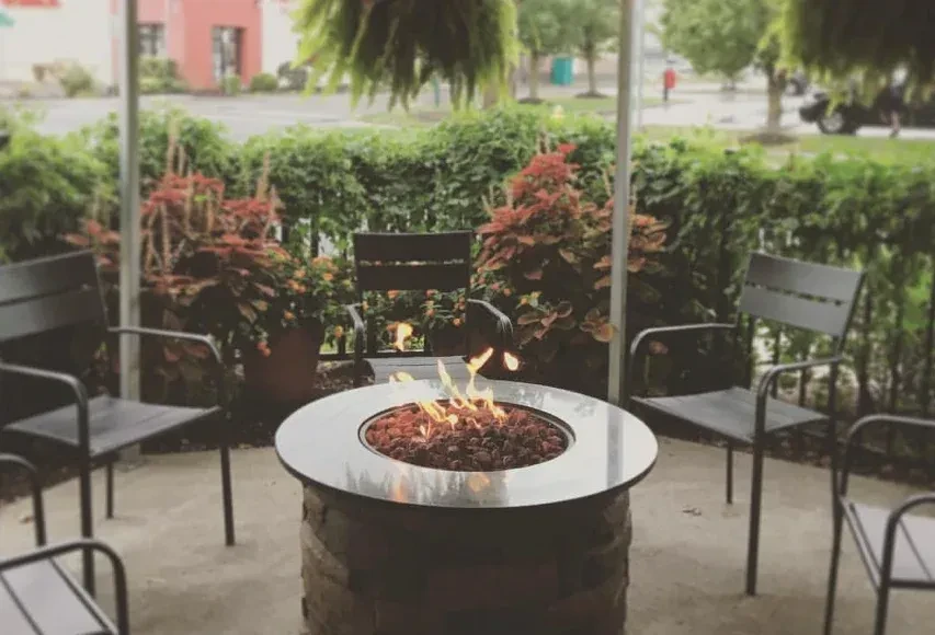 Burning fire pit on an outdoor patio, surrounded by chairs and foliage.