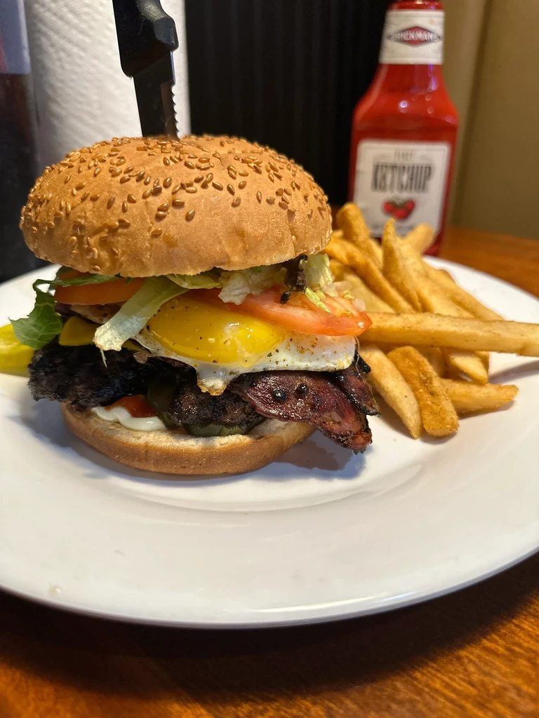 Close-up: tall burger with fried egg, bacon, cheese, knife, and fries.