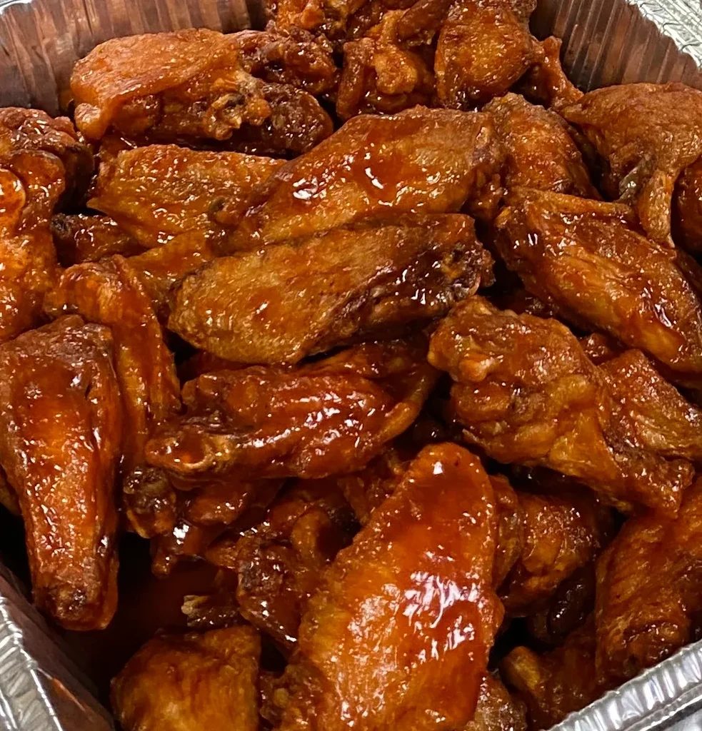 Piled high chicken wings in a foil pan with glossy reddish-brown sauce.