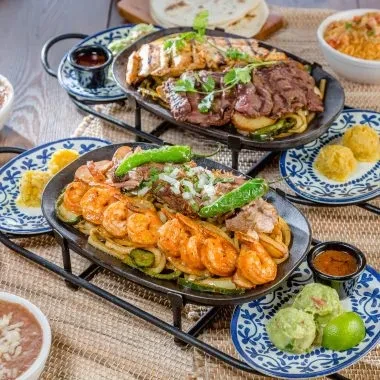 Two sizzling platters of grilled meats, seafood, vegetables, and Mexican sides.