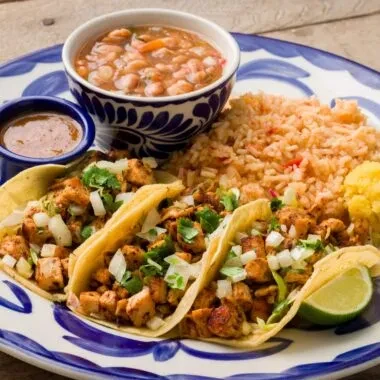 Three Mexican tacos, rice, beans, lime, on a patterned plate.