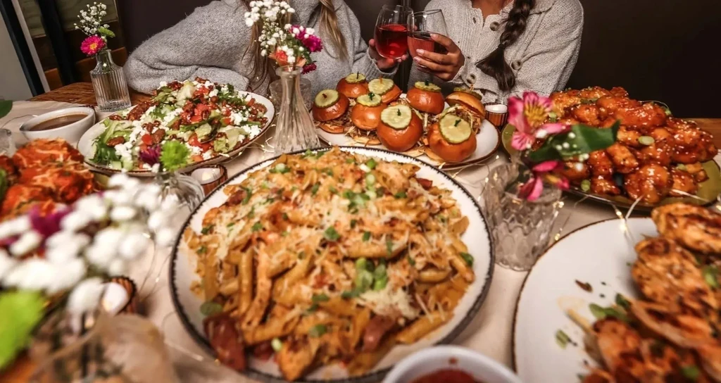 Dining table overflowing with various dishes; two hands toast with wine.