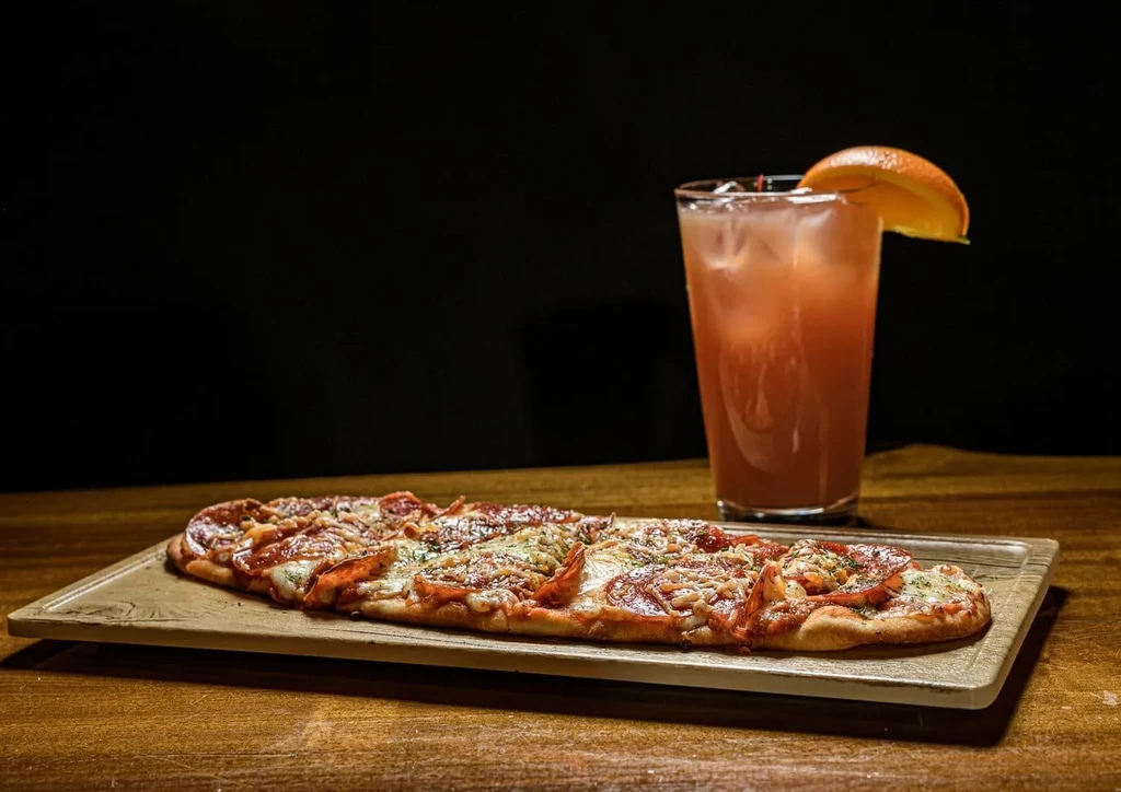 Rectangular pepperoni flatbread pizza and amber cocktail on a dark wooden table.